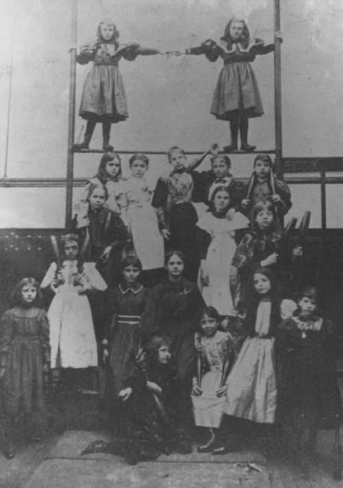 A historic black-and-white photo of a group of girls posing in two tiers, dressed in various vintage dresses.