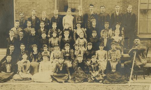 A large group photo of students and a teacher, dressed in late 19th-century clothing, posed outdoors in front of a building.