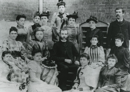 A historical black-and-white photo of a group of 15 men and women, dressed in 19th-century clothing, posing together.