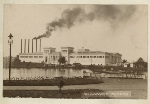 Historic photo of a large machinery building by a lake, with smoke rising from tall chimneys in the background.
