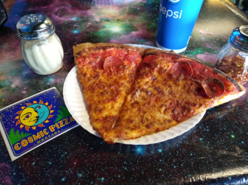Two slices of pepperoni pizza on a paper plate, with a soda and condiments on a colorful, space-themed table.