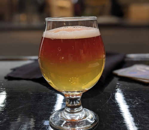 A glass of beer with a gradient from amber to golden yellow, sitting on a dark surface.