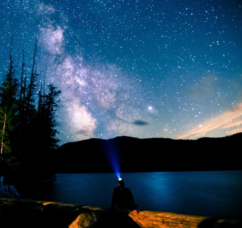 A person sits by a lake at night, gazing at a starry sky with the Milky Way, illuminated by a headlamp.