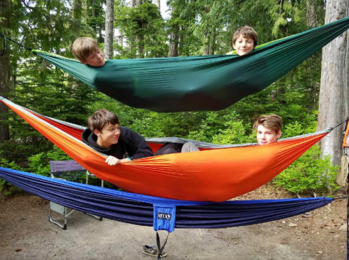 Four boys playfully lounging in colorful hammocks strung between trees in a forested campsite.