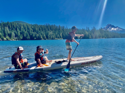 Three children paddleboarding on a clear lake, surrounded by trees and mountains under a bright blue sky.
