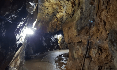 A dimly lit cave with rocky walls and a winding path, illuminated by lights along the route.