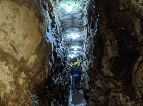 A narrow, illuminated tunnel with rocky walls, featuring a person wearing a yellow helmet walking through it.