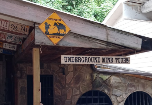Sign for "Underground Mine Tours" under a prospector crossing warning, with a rustic wooden structure in the background.