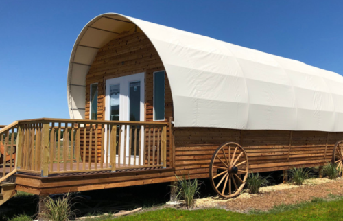 A wooden wagon-style structure with a curved white roof, featuring a porch and surrounded by greenery.