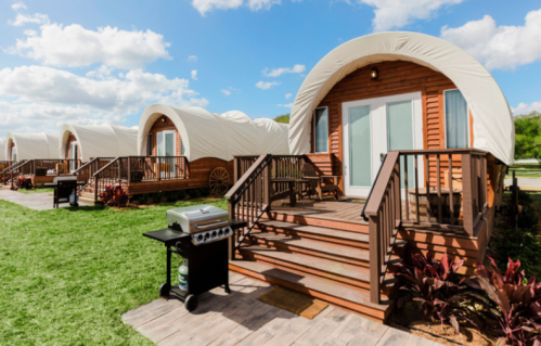 Four wooden cabins with curved roofs, each with a porch and grill, set on a grassy area under a blue sky.