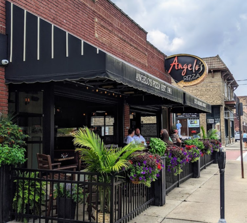 Exterior of Angelo's Pizza restaurant with outdoor seating, plants, and a sign, set against a cloudy sky.