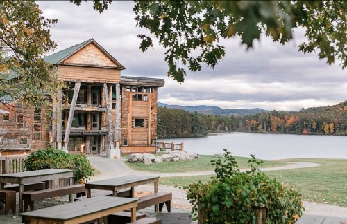 A rustic wooden building by a lake, surrounded by trees with autumn foliage under a cloudy sky.