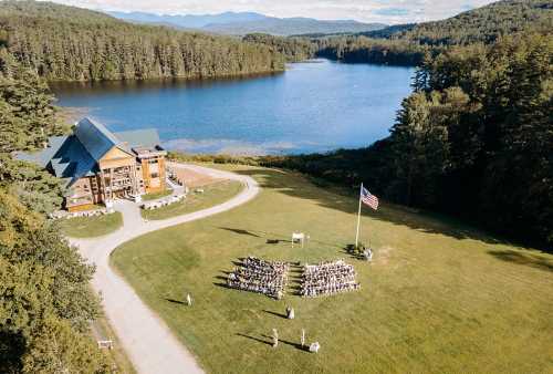 Aerial view of a lakeside venue with a large gathering on the lawn, surrounded by trees and mountains in the background.