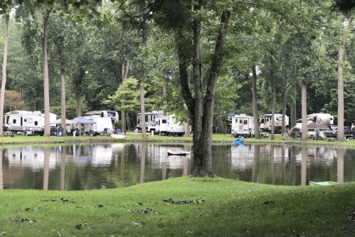 A serene campsite with RVs parked near a calm pond, surrounded by lush green trees.