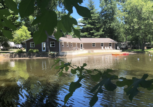 A serene lakeside view of a brown house with a deck, surrounded by trees and a calm water surface.