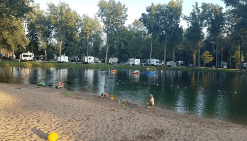 A serene lake surrounded by trees, with campers in the background and a child playing in the sand by the water's edge.