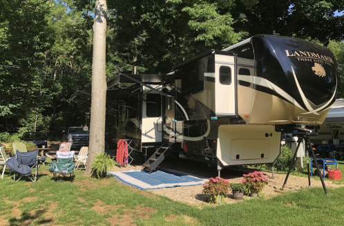 A large RV parked in a wooded area, with outdoor chairs and a blue mat set up nearby, surrounded by greenery.