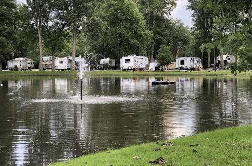 A serene pond with a fountain, surrounded by green grass and RVs parked in the background under a cloudy sky.