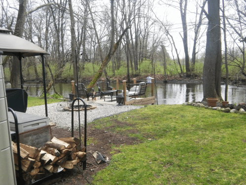 A serene riverside scene with a fire pit area, chairs, and a stack of firewood surrounded by trees and greenery.