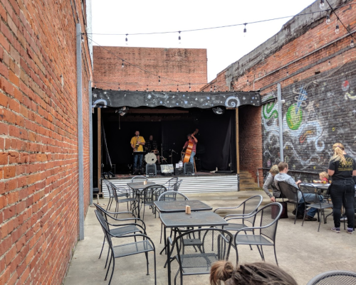 Outdoor venue with a stage featuring musicians, surrounded by brick walls and tables for patrons.