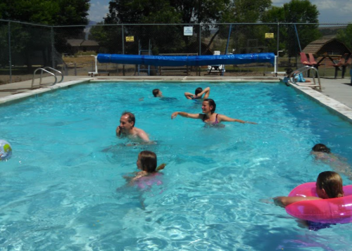 A group of children and adults swimming and playing in a bright blue pool on a sunny day.