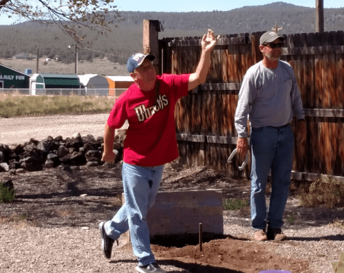 Two men outdoors; one in a red shirt throws a horseshoe while the other watches, with mountains in the background.