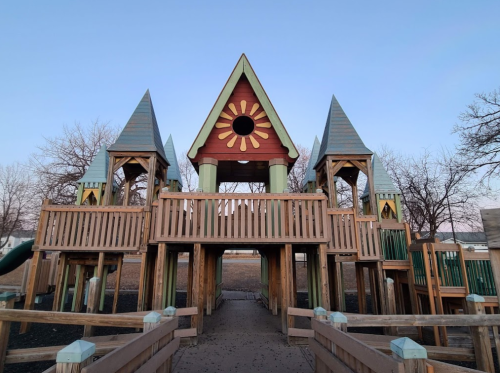 Colorful wooden playground structure with two towers and a sun design, surrounded by trees and a clear sky.