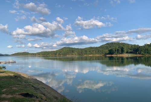 A serene lake reflecting clouds and trees, surrounded by hills under a clear blue sky.