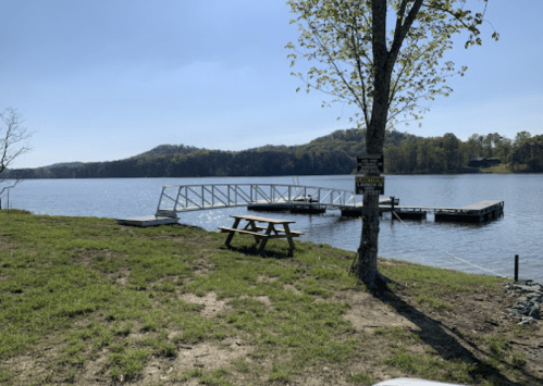 A serene lakeside view featuring a dock, picnic table, and lush green hills in the background under a clear blue sky.