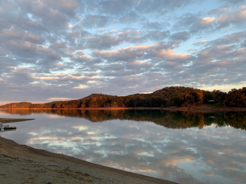 A serene lake at sunset, reflecting colorful clouds and trees along the shore under a peaceful sky.
