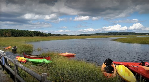 Colorful kayaks lined up by a serene river, surrounded by lush green grass and a cloudy sky.