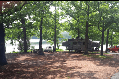 A campsite with a trailer and tent surrounded by trees, near a lake on a sunny day.