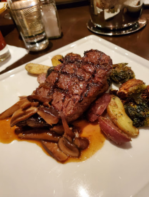 A beautifully plated steak with grilled vegetables and mushrooms, drizzled with sauce on a white dish.