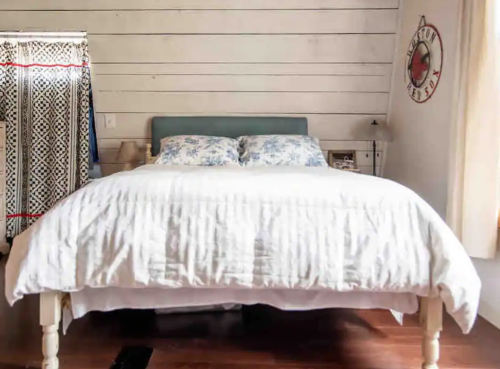 Cozy bedroom featuring a light blue headboard, white bedding, and a patterned curtain against a wooden wall.