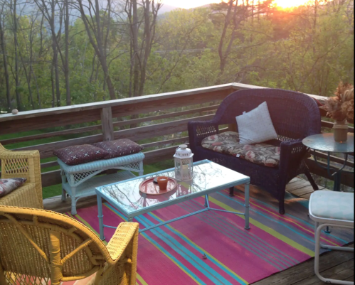 A cozy outdoor patio with colorful furniture, a glass table, and a sunset view through trees.