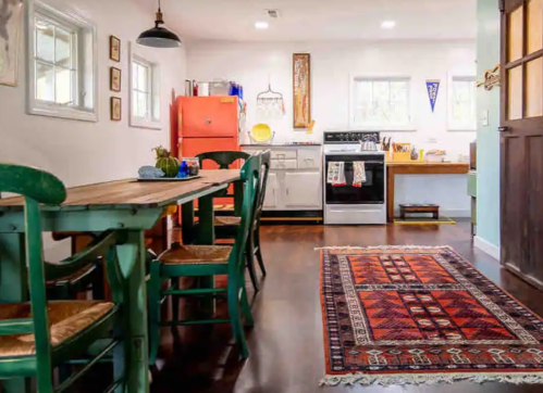 Bright kitchen with a red fridge, wooden dining table, green chairs, and a colorful rug on dark hardwood floors.