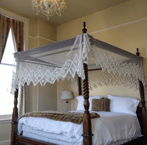 A vintage four-poster bed with a lace canopy in a softly lit room featuring warm yellow walls and elegant decor.