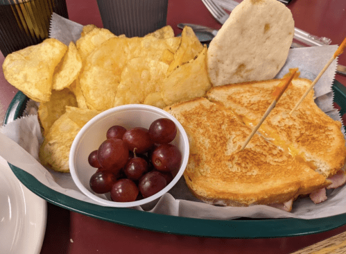 A basket with a grilled cheese sandwich, potato chips, and a cup of grapes.