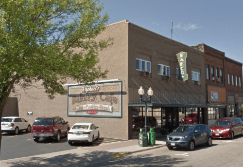 A bakery and café building with a large sign, surrounded by parked cars and trees on a sunny day.