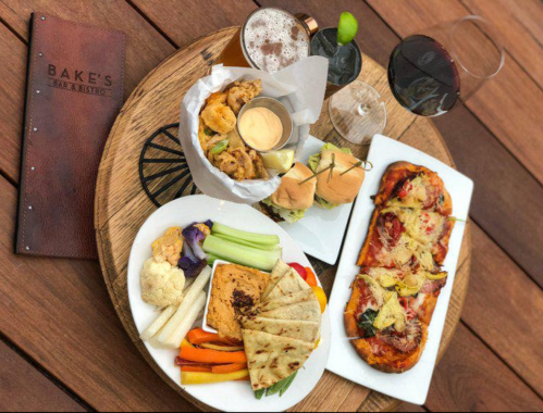 A wooden platter with various dishes: sliders, flatbread, veggies, and drinks, alongside a menu from Bake's Bar & Bistro.