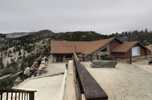 A rustic building with a sloped roof, surrounded by mountains and trees, with people gathered outside on a gravel path.