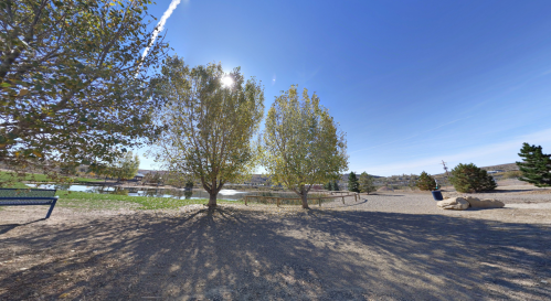 A sunny park scene with two trees, a gravel path, and a pond in the background, surrounded by green grass and distant hills.