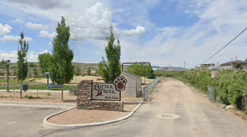 Sign for Bitter Creek Bark Park with a dirt path and green trees under a cloudy sky.