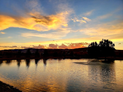 A serene sunset over a calm lake, with colorful clouds reflecting on the water and silhouettes of trees in the foreground.