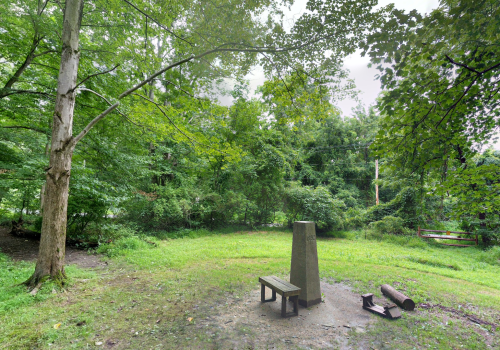 A serene green park with a stone monument, a wooden bench, and logs on the ground, surrounded by lush trees.