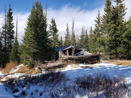 A dilapidated cabin surrounded by snow and evergreen trees under a partly cloudy sky.