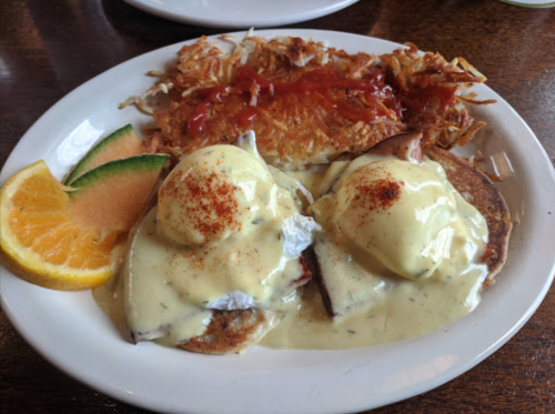 A plate of eggs Benedict with hollandaise sauce, served with hash browns and garnished with orange and lime slices.