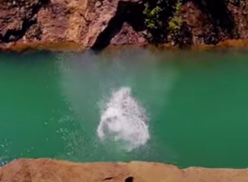 A splash of water erupts from a turquoise pool as someone dives in from a rocky edge.