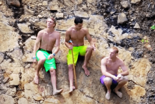 Three young men sit on a rocky surface, wearing bright swim trunks, enjoying a sunny day outdoors.