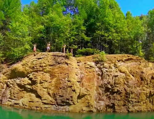 A group of people stands on a rocky ledge by a clear blue lake, surrounded by lush green trees under a bright sky.